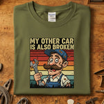 My Other Car Is Also Broken T-Skjorte med morsom mekanikerillustrasjon på olivengrønn bakgrunn.